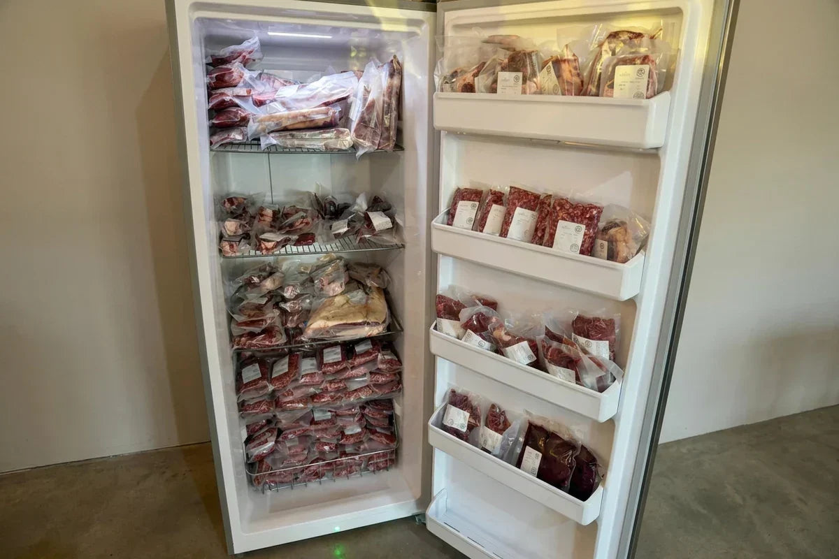 Open upright freezer filled with vacuum-sealed raw meat packages on wire shelves.